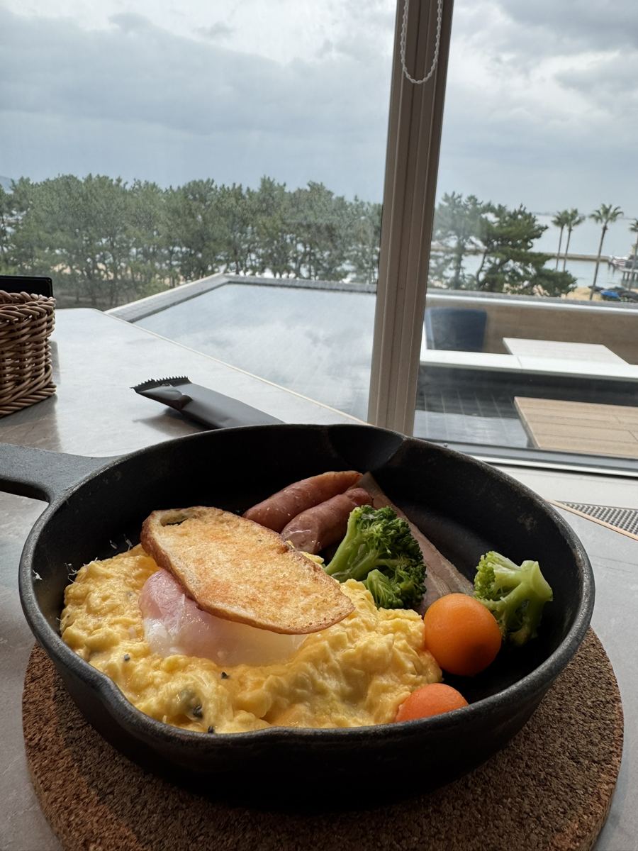 Scrambled eggs, sausages, broccoli and toast in a cast-iron skillet by a window with a sea view