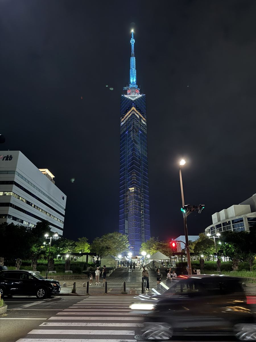 Fukuoka Tower lit up blue at night, seen from a street crossing