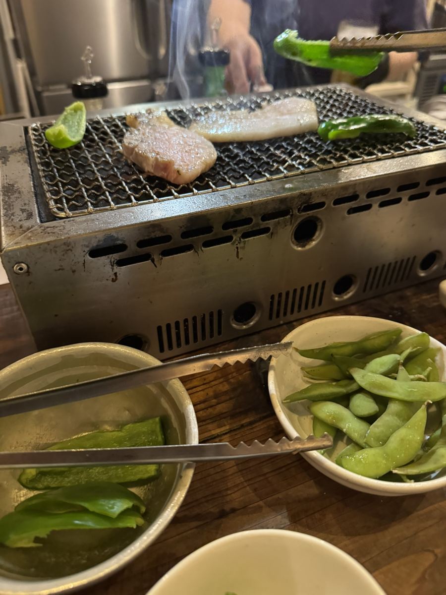 A tabletop charcoal grill with pork belly and green peppers, bowls of edamame and peppers in front