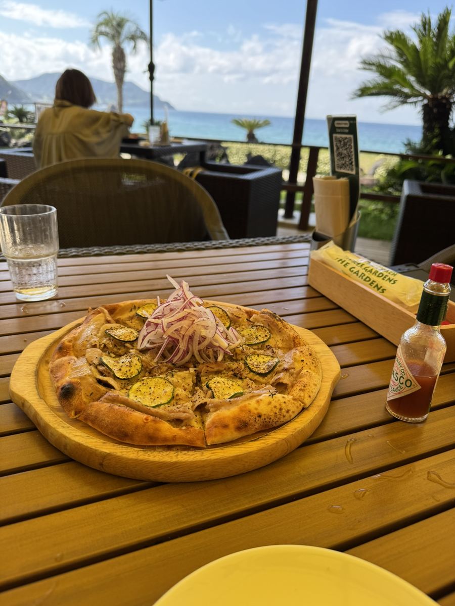 A pizza with zucchini and red onion on a wooden table at a beachside café, with palm trees and the sea behind