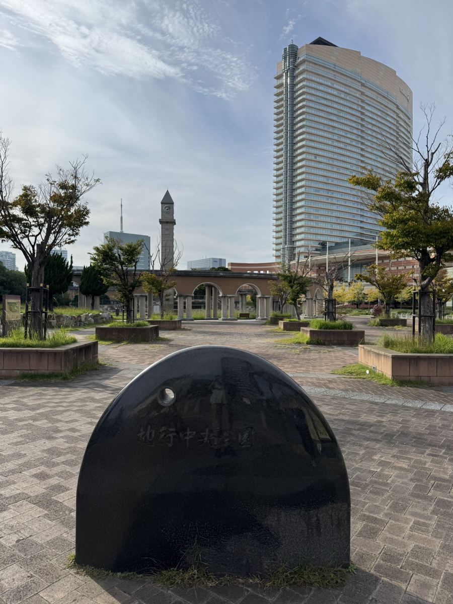 A park with a stone marker, a brick colonnade with a clock tower and a tall curved hotel tower behind it