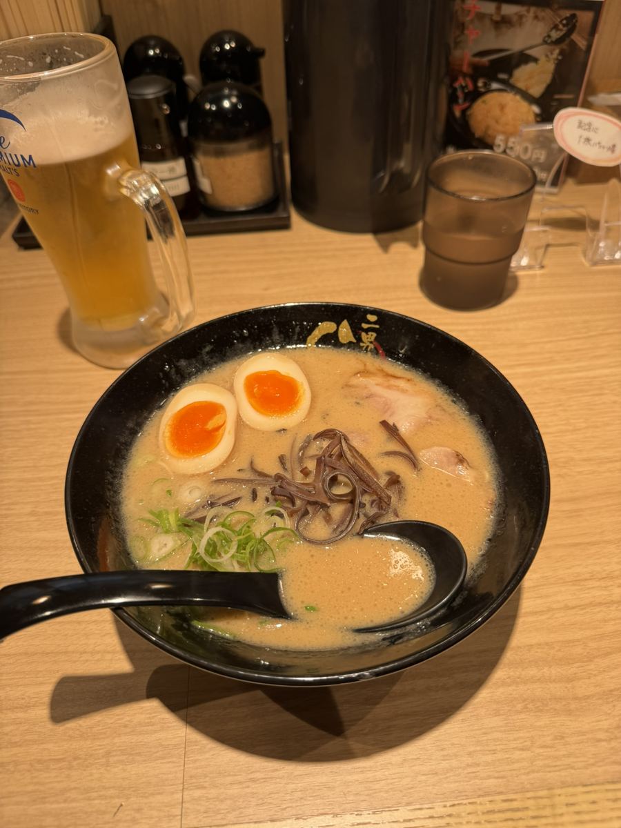A bowl of tonkotsu ramen with a soft-boiled egg, green onions and wood ear mushrooms, next to a glass of beer