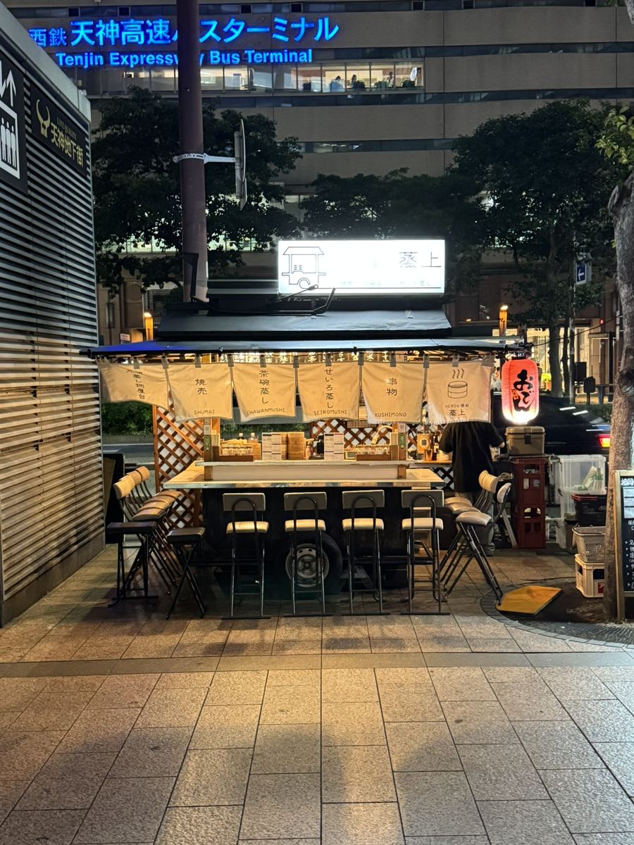 A yatai food stall with white curtains and empty stools in front of the Tenjin Expressway Bus Terminal at night